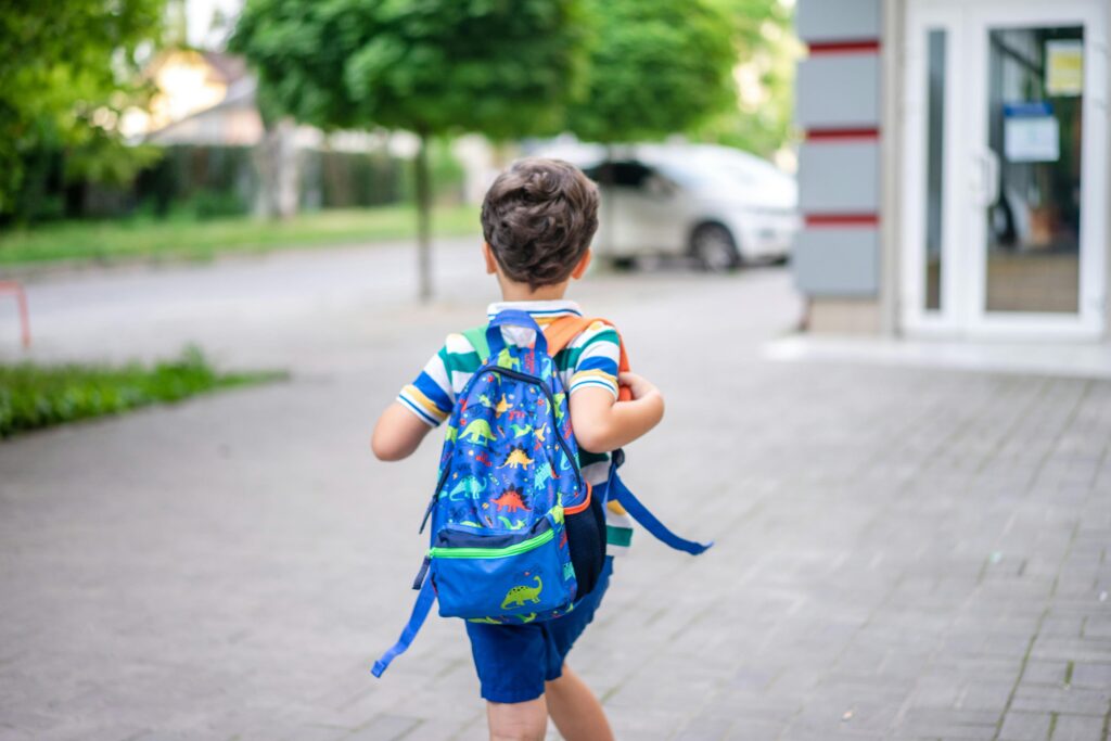 Back view of a child with a colorful dinosaur backpack walking on a sidewalk in a suburban area.