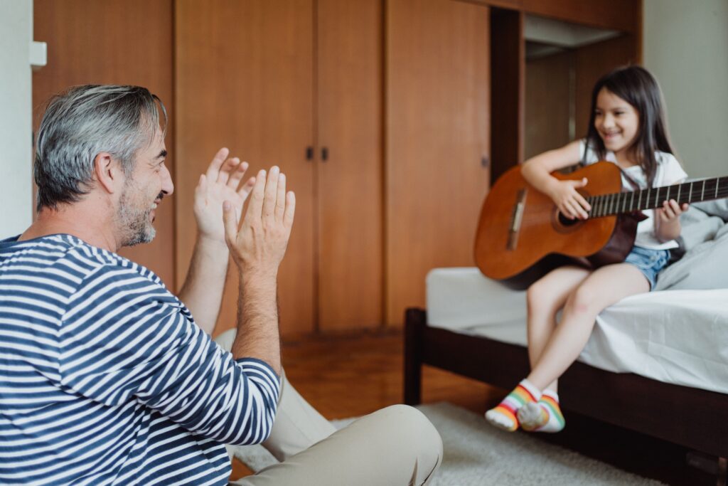 A joyful father and daughter share a musical moment as she plays guitar in the room.