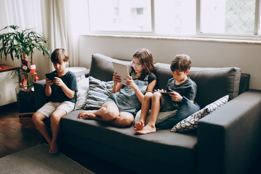 Three children on a gray sofa engrossed in digital devices, indoors.