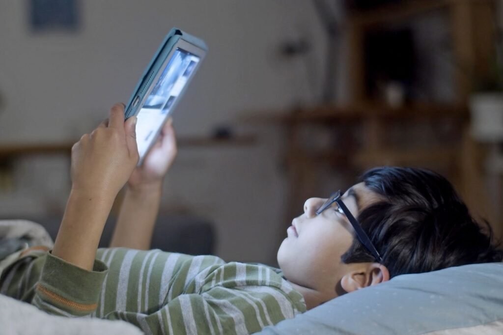 A young boy wearing glasses uses a tablet while lying in bed at night, creating a cozy and focused atmosphere.