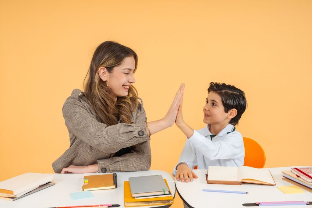 pexels-photo-35745342-35745342 Teacher and student sharing a high-five over a desk in a bright classroom, fostering a positive learning environment.