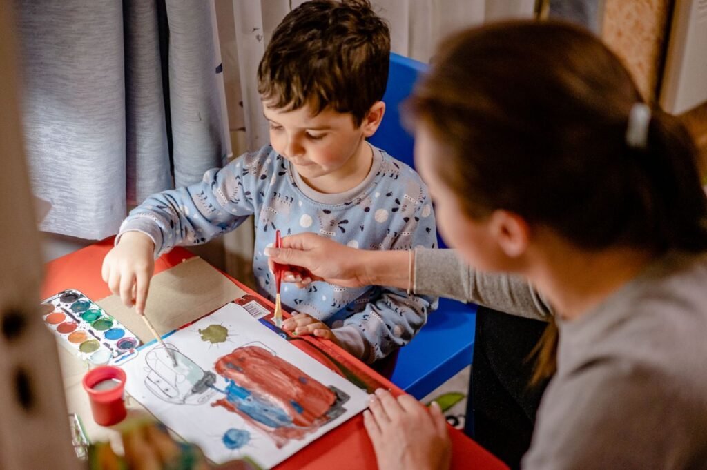 pexels-photo-7676908-7676908 High angle of mum with paintbrush helping son in drawing picture with watercolor paints in kids bedroom