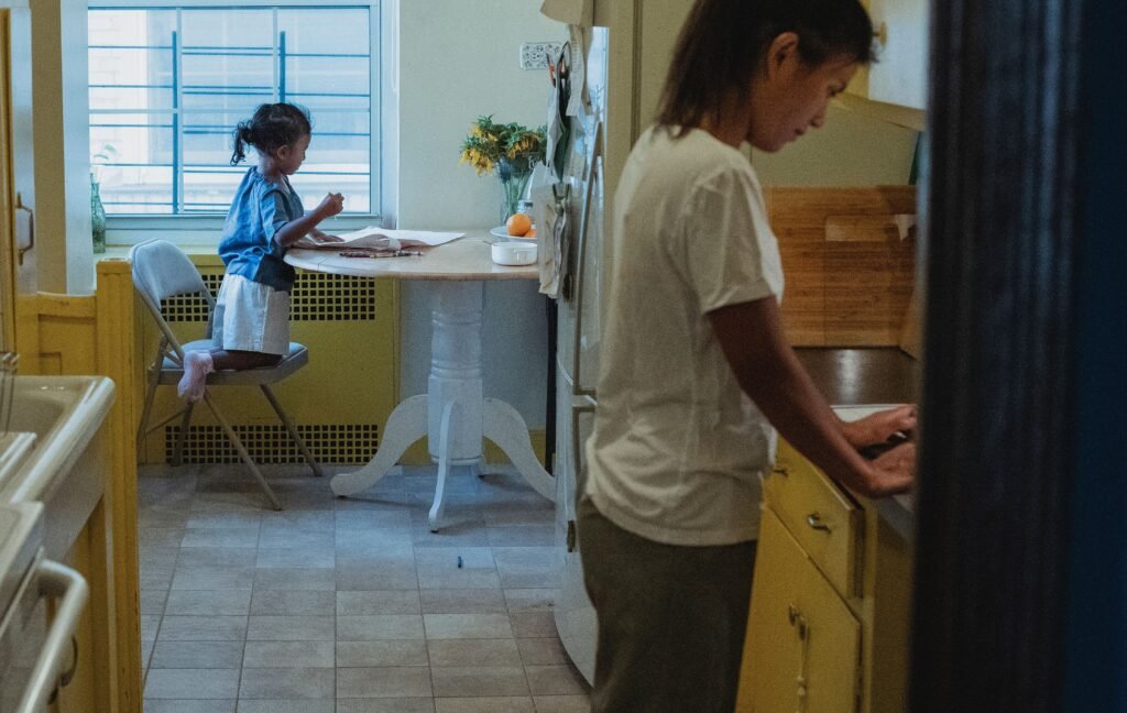 Side view of busy ethnic woman standing near cupboards on kitchen with little girl drawing at table during weekend at home