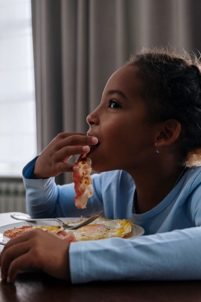 Young girl happily eating bacon and eggs at home during breakfast time.